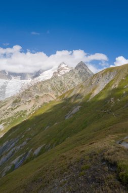 Dik yeşil tepeler, Buzul de Pre de Bar ve Mont Dolent yakınlarında dağınık bulutlarla canlı mavi bir gökyüzünün altında dramatik bir buzulun ve sivri bir dağın zirvesine çıkar..