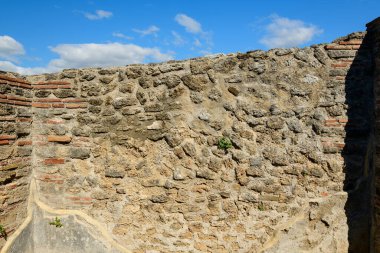 İtalya 'nın Paestum kentindeki eski harabelerden kalma yıpranmış taş ve tuğla bir duvara yakın çekim. Dağınık bulutlarla dolu canlı mavi gökyüzünün altında gün ışığıyla aydınlatılıyor. Taş ustalığı tarihin katmanlarını gözler önüne seriyor ve çatlaklardaki bitkilerin büyümesini inceliyor..