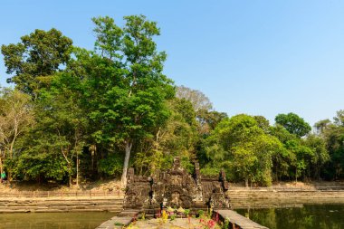 Antik taş tapınak, Angkor bölgesindeki Neak Pean Tapınağı 'nda yemyeşil ağaçlar ve açık mavi gökyüzü ile çevrili dikdörtgen bir göletin merkezinde yer alır. Yıpranmış oymalar ve sakin su, tropikal suların ortasında sakin bir atmosferi çağrıştırıyor.