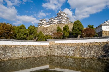 Himeji Kaleleri ikonik beyaz yapı katmanlı taş surların üzerinde yükselir ve yeşil ve sonbahar ağaçlarıyla çevrili sakin bir hendek dağınık bulutlarla kaplı parlak bir gökyüzü altında. Güneş ışığı, tarihi kalenin ve dingin suyun dokusunu vurguluyor.