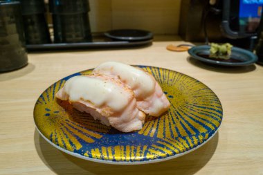 Eritilmiş peynirle kaplanmış iki parça kurutulmuş somon nigiri suşisi, çağdaş bir Osaka suşi restoranında soluk ahşap bir tezgahın üzerine kurulmuş mavi ve sarı desenli bir tabakta sunuluyor. Görüntü kremsi dokular ve sıcak, davetkar