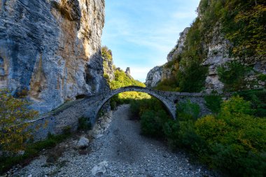 Arched stone bridge spans a dry rocky riverbed between towering limestone cliffs and dense autumn foliage in the Vikos Gorge region of Epirus, Greece. Soft daylight reveals textured masonry and vibrant natural surroundings in this dramatic mountain