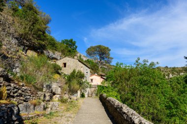 Dar bir taş geçit, Navacelles 'deki kayalık tepelerin ve yemyeşil yeşilliklerin arasına kurulmuş geleneksel bir taş eve açılıyor. Parlak güneş ışığı ve berrak mavi gökyüzü, duvarcılık ve doğal manzaranın dokusunu vurgular..