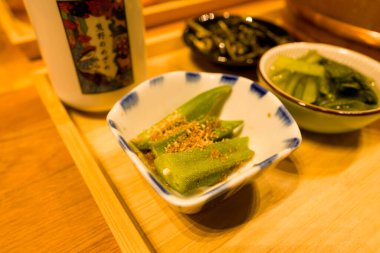 Dilimlenmiş bamya dilimlenmiş susam tohumları, ahşap bir tepside, mavi kenarlı bir seramik kasede sunulur. Yanında Japon yemek takımında, çeşitli sebze tabakları bulunur..