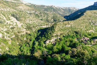 Navacelles Sirki 'nin dramatik kireçtaşı tepelerinin dibinde yoğun yeşil ormanlarla çevrili küçük bir taş ev kümesi bulunur. Parlak güneş ışığı engebeli kanyon manzarasında kıtır kıtır gölgeler bırakır.