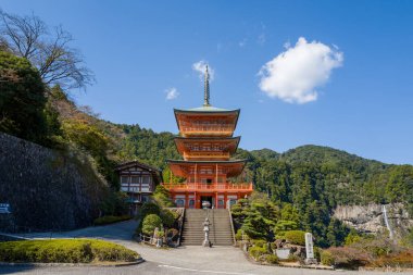 Japonya 'nın Wakayama eyaletinin Kumano Nachi Taisha kentindeki açık mavi gökyüzünün altında kayalık uçurumlardan dökülen uzak bir şelaleyle, üç katlı yemyeşil tepelerin eteklerinde çarpıcı bir şelale duruyor. Parlak güneş ışığı canlı renkleri ve karmaşıklığı vurgular