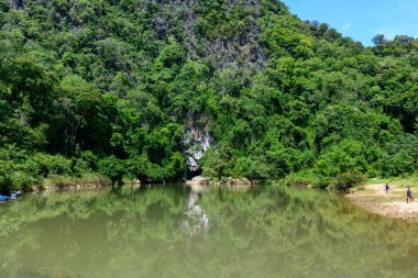 Yoğun tropikal orman battaniyeleri sakin bir nehrin üzerinde engebeli kireçtaşı uçurumlar oluştururken, Konglor Mağarası 'nın girişi su kenarında görülebiliyor. Sakin yüzey, Laos 'un tipik yeşil ve dramatik kaya oluşumlarını yansıtır..