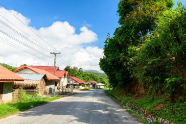 Kırmızı metal çatıları olan evlerle dolu sessiz bir köy yolu, yoğun yeşil bitki örtüsü ve yüksek ağaçlarla birlikte kuzey Laos tepelerinde dağınık bulutlarla dolu canlı mavi bir gökyüzünün altında uzanır. Sahne, kırsal mimariyi ve genel gücü ön plana çıkarıyor.
