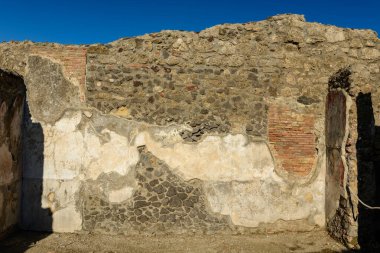 A weathered stone and brick wall stands under clear blue sky in the ruins of Pompeii, Italy. The surface reveals layers of ancient masonry, faded plaster, and volcanic rock, capturing the enduring character of Roman architecture.