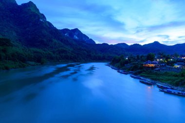 Loş alacakaranlık Nam Ou Nehri 'ni ve Laos' un Nong Khiaw köyünü aydınlık evler ve sakin nehir kıyısında yüzen teknelerle kaplar. Bereketli yeşil dağlar ve canlı mavi bir gökyüzü bu manzaralı nehir kıyısında sakin bir manzara yaratır..