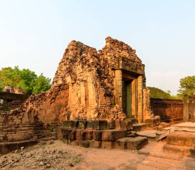 Sıcak güneş ışığı, Kamboçya 'daki Pre Rup tapınağının yıpranmış kırmızı tuğlalarını ve yan duvarlarını yıkıyor. Sahnede aşınmış duvarcılık, karmaşık taş oymacılığı ve açık bir gökyüzü altında yeşillikle çerçevelenmiş sükunet ortamı yer alıyor..