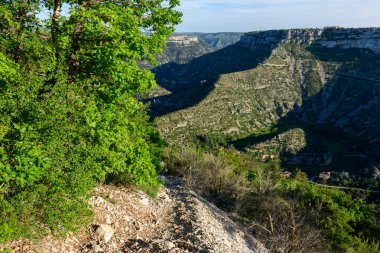 Bereketli yeşil yaprakların yanına uzanan engebeli bir patika derin kireçtaşı uçurumlarını ve Navacelles Sirki 'nin dolambaçlı vadisini kaplıyor. Parlak güneş ışığı, kaya ve bitki örtüsünün dokusunu vurgular.