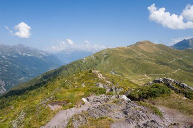Rocky mountain ridge overlooks rolling green slopes crisscrossed by winding paths under a clear blue sky in the Aiguillettes des Posettes region.