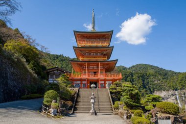 Manikürlü yeşillik ve Japonya 'nın Wakayama eyaletindeki Kumano Nachi Taisha ormanlı tepelerle çevrili taştan basamakların üzerinden canlı turuncu pagoda yükselir. Parlak güneş ışığı ve berrak mavi bir gökyüzü uzak bir şelale ile huzurlu ve neşelendirici bir atmosfer yaratır..