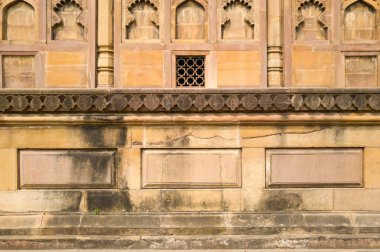 Prayagraj 'daki Khusro Bagh' da süslü oyma paneller ve geometrik kafes işi içeren yıpranmış kumtaşı duvar. Güneş ışığı, tarihi anıtın eski dokularını ve ince renk değişimlerini vurguluyor.