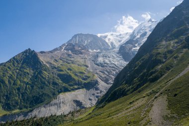 Yüksek yeşil dağlar arasındaki dik kayalık yamaçlardan aşağı süzülen bir buzulun dramatik manzarası. Kar yığınları ve açık mavi bir gökyüzü..