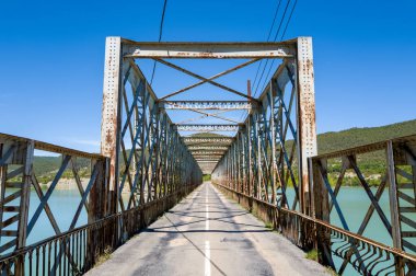 Paslanmış kirişleri olan yıpranmış metal bir kiriş köprüsü, açık mavi-yeşil rezervuar suyu boyunca uzanır. Etrafı orman tepeleri ve Las Penas 'ta canlı mavi bir gökyüzü ile çevrilidir. Güçlü geometrik çizgiler ve açık yollar derinlik ve macera hissi yaratır..