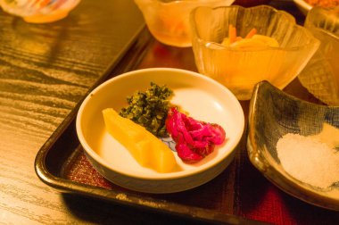 Japon salamura sebzelerini küçük bir tabakta, Hongu, Japonya 'da sıcak ortam aydınlatması altında sunulmuştu. Sahne canlı renkleri ve geleneksel dokuları vurguluyor.