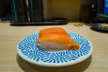 Modern bir Osaka suşi restoranında soluk ahşap bir tezgahın üzerine kurulmuş koyu mavi çiçekli beyaz bir tabakta duran iki parça taze somon nigiri suşi. Sahne canlı rengi ve minimalist sunumu vurguluyor.