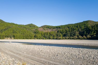 Wakayama, Hongu 'da açık mavi bir gökyüzünün altında yemyeşil ormanlık tepelerin yanında akan dar bir dereyle geniş kayalık nehir yatağı. Manzara açık ve güneşli. Huzur ve dokunulmamış doğa hissi uyandırıyor..