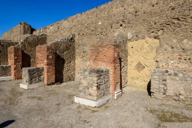 Yıpranmış tuğla sütunlar ve volkanik taş sayaçlar İtalya 'nın Pompeii kentindeki yerleşim yerlerinde antik taş duvarlara karşı dururlar. Parlak güneş ışığı ve parlak mavi gökyüzü bu arkeolojik yüzeyin geometrik dokularını ve tarihi yüzeylerini vurgular.