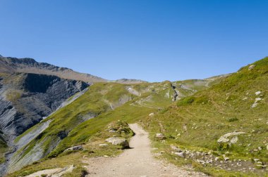 Col de la Seigne yakınlarındaki İtalyan vadisinde, bulutsuz mavi bir gökyüzünün altında yeşil alp tepeleri ve kayalık çıkıntılar arasında bir toprak yürüyüş yolu esiyor. Manzara açık ve güneşli, engebeli dokuları ve bir huzur hissi var..