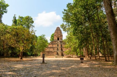 Baksei Chamkrong 'un antik tuğla piramidi Angkor, Kamboçya yakınlarındaki ağaçlık alanının sonunda yükseliyor. Sıcak gün ışığı, dağınık bulutlu mavi gökyüzünün altında yoğun yeşil yapraklarla çevrili yıpranmış basamakları ve dokulu duvarları vurgular..