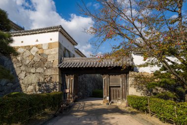 Yıpranmış ahşap I No Mon kapısı, Himeji Kalesi 'nde taş duvarların ve kiremitli çatıların altında duruyor, yapraklı çitlerle çerçevelenmiş ve dağınık mavi gökyüzü altında seyrek sonbahar yeşillikleri..