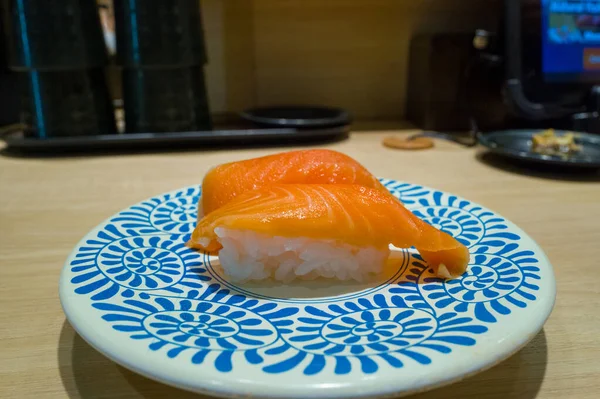 Çağdaş bir Osaka suşi restoranının açık ahşap tezgahının üzerinde mavi ve beyaz çiçekli bir tabakta duran iki parça taze somon nigiri suşisi. Sahnede canlı renkler ve temiz ve minimal bir sunum yer alıyor..