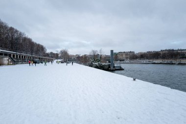 Paris, Fransa - 7 Ocak 2026: Seine nehrinin kenarındaki yapraksız ağaçlar, sokak lambaları ve arka planda bulutlu bir gökyüzünün altında görünen Haussmann tarzı apartmanlar bulunan geniş bir rıhtımda birkaç kişi yürüyüp koşu yapıyor..
