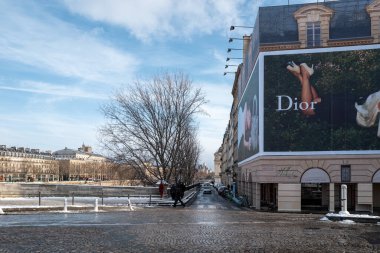 Paris, Fransa - 7 Ocak 2026: Büyük bir Dior reklamı Seine Nehri yakınlarındaki bir cadde köşesinde yayalar, yapraksız ağaçlar ve parçalı bulutlu gökyüzü altında görünen tarihi binaların bulunduğu bir binanın ön yüzünü kapsıyor..