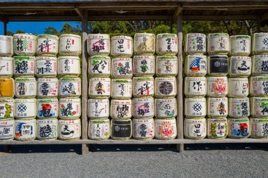 Ise, Japonya - 29 Ekim 2023: Japon yazıları ve renkli tasarımları olan birçok sıra saki fıçısı büyük ahşap bir rafta, yerde çakıllar ve arka planda görünen ağaçlar üzerinde düzenlenmiştir..