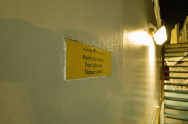 Bilinmeyen konum - 13 Eylül 2023: Görüntü, arka planda ışıklı merdivenlerle gece feribotun dış duvarında birden fazla dilde Kaygan Güverte yazan sarı bir uyarı işareti gösteriyor..