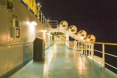 Bilinmeyen konum - 13 Eylül 2023: Görüntü, yukarıda monte edilmiş cankurtaran botları, aydınlatılmış geçitler, güvenlik korkulukları ve yapay aydınlatma altında bir çöp tenekesinin bulunduğu gece bir feribotun dış güvertesini gösteriyor..