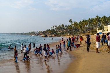 Kovalam, Hindistan - 1 Mart 2023: Grove Beach 'in kumlu kıyı şeridi boyunca oynayan ve ayakta duran çocuk ve yetişkin grupları, palmiye ağaçları, binalar ve parçalı bulutlu bir gökyüzünün altında görünen deniz.