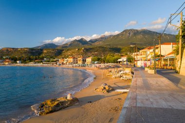 Stoupa, Yunanistan - 12 Eylül 2024: Görüntü, güneş pansiyonları ve şemsiyeli kumlu bir plajı, deniz kenarındaki binalarla çevrili bir gezinti alanı ve gün batımında görülebilen dağları gösteriyor.