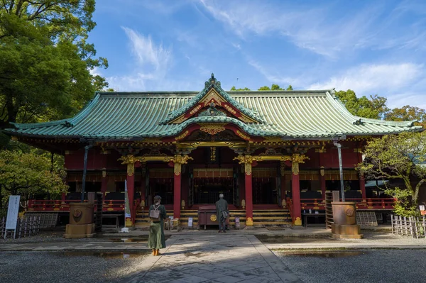 Tokyo, Japonya - 30 Ekim 2024: Görüntü, yeşil kiremit çatılı ve mavi gökyüzü altında dekoratif mimari ayrıntılara sahip geleneksel kırmızı ve altın ahşap bir yapı olan Nezu Tapınağı 'nın ana ibadet salonunun önünde duran iki kişiyi gösteriyor..