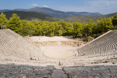 Epidaurus, Yunanistan - 2 Eylül 2024: Antik Asclepius Sığınağı 'ndaki yarı dairesel tiyatronun üst taş oturma yerinden, orkestra alanını, dağınık ziyaretçileri, çam ağaçlarını çevreleyen ve kısmen uzak dağları gösteren