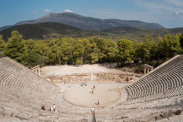 Epidaurus, Yunanistan - 2 Eylül 2024: Asclepius Sığınağı 'ndaki antik yarı dairesel taş tiyatronun geniş görünümü, yıpranmış taş koltuklar, orkestra alanındaki ziyaretçiler, etrafındaki çam ağaçları ve uzak dağları gösteriyor