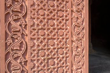 Agra, Hindistan - 11 Mart 2024: Görüntü Agra Kalesi 'ndeki Jahangir Sarayı' nın duvarına kırmızı kumtaşına oyulmuş ve güneş ışığıyla aydınlatılan karmaşık çiçek ve geometrik desenlerin yakın çekimini gösteriyor.