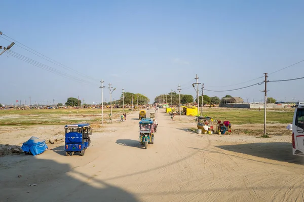 Prayagraj, Hindistan - 21 Mart 2024: Görüntü, elektrik direkleriyle, oto çekçekçekçeklerle, yol kenarı tezgahlarıyla, dağınık insanlarla ve açık bir gökyüzü altında gün ışığında açık bir zemin altında uzanan kumlu bir yol gösteriyor.