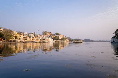 Udaipur, Hindistan - 21 Mart 2022: Görüntü, şehir sarayı ve Pichola Gölü kıyısı boyunca uzanan tarihi sarı ve beyaz binaları, berrak bir gökyüzünün altındaki sakin suda yansımalarını gösteriyor..