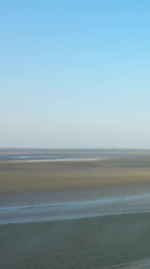 Fransa, Le Mont-Saint-Michel 'de açık mavi gökyüzünün altında kumlu gelgit düzlükleri ve sığ su manzarası. Doğal dokuları ve yumuşak ışıkla huzurlu kıyı manzarası.