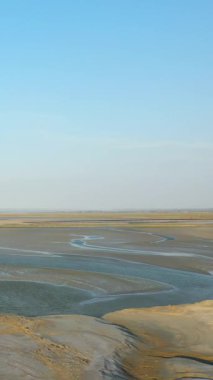 Fransa 'nın Le Mont-Saint-Michel şehrinde açık mavi gökyüzünün altında dalgalı su kanalları olan geniş gelgit düzlükleri. Kum ve sığ su desenleriyle sakin doğal manzara.