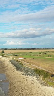 Taşlı bir plajın, açık arazilerin ve parçalı bulutlu bir gökyüzünün altında küçük beyaz bir evin bulunduğu kırsal bir sahil manzarası. Barışçıl kırsal kesim doğal dokuları ve geniş manzarayı ön plana çıkarıyor.