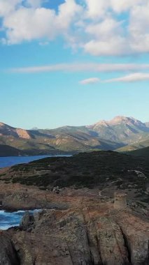 Engebeli kıyı şeridi manzarası, tarihi taş kule ve Cargese, Korsika yakınlarında bulutlu mavi bir gökyüzünün altında dağ arkaplanı..