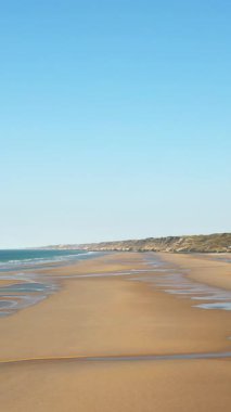 Fransa, Normandiya 'daki Omaha Sahili' nin geniş manzarası, altın kum, hafif dalgalar ve açık mavi gökyüzü. Huzurlu kıyı manzarası seyahat ve doğa kavramları için ideal..