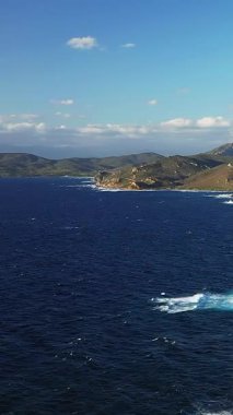 Les Iles Sanguinaires 'teki engebeli kıyı şeridi ve derin mavi denizin manzarası berrak bir gökyüzünün altında. Görüntü kayalık kıyıya çarpan dalgaları yakalıyor. Arka planda uzak tepeler var..