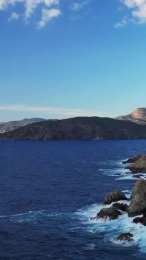 Dalgalar, Corsica, Cargese yakınlarındaki açık mavi gökyüzünün altında uzak tepeleri olan engebeli bir sahil şeridi boyunca kayalara çarpar..