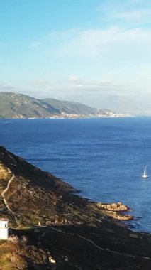 Mavi denize bakan kayalık bir kıyı şeridindeki deniz fenerinin manzarası, uzak dağlar ve Korsika 'daki Les Iles Sanguinaires yakınlarında bir yelkenli..